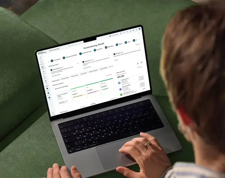 QuickBooks Online dashboard on a laptop showing the Business Feed, Payments Agent, Accounting Agent, bank account balances, and Business at a Glance summary